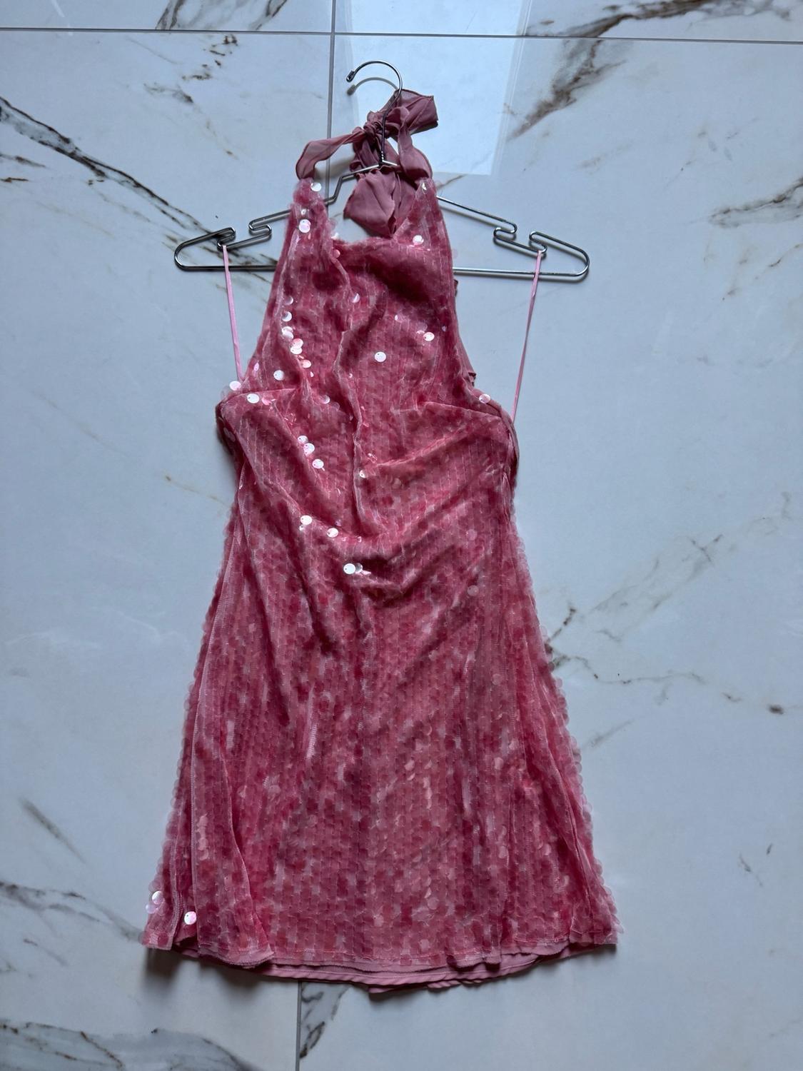 Κοντό ροζ φόρεμα με παγιέτες και λαιμόκοψη halter, κρεμασμένο σε κρεμάστρα σε φόντο λευκού μαρμάρου.