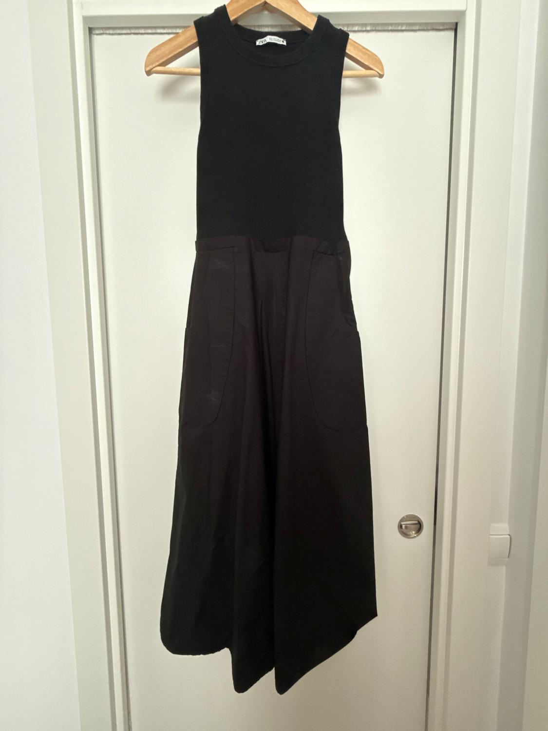 Black dress hanging on a wooden hanger in front of a white door. ZARA label. Sleeveless design, fitted bodice and flared skirt with pockets.