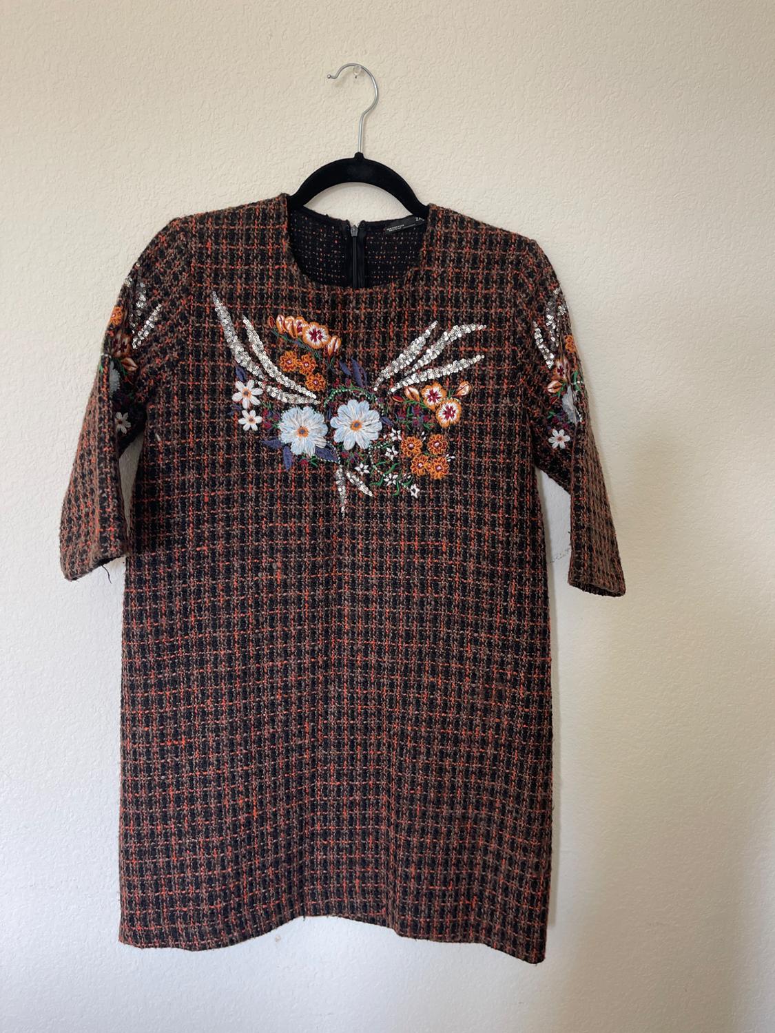 Short-sleeved dress with a dark checked pattern and colourful floral embroidery on the chest, hanging on a black hanger.