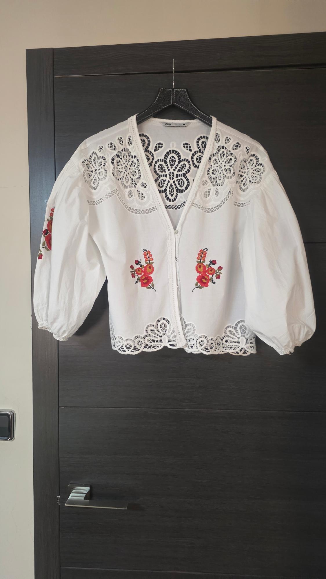 White blouse with red floral embroidery and lace, hung on a dark door. Puffed sleeves and open-front design.