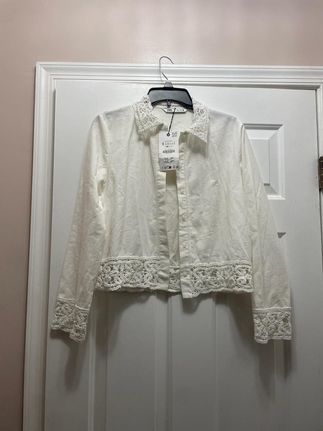 White long-sleeved shirt with lace on the collar, cuffs and hem, hanging on a white door. Tag with the text zara and S.