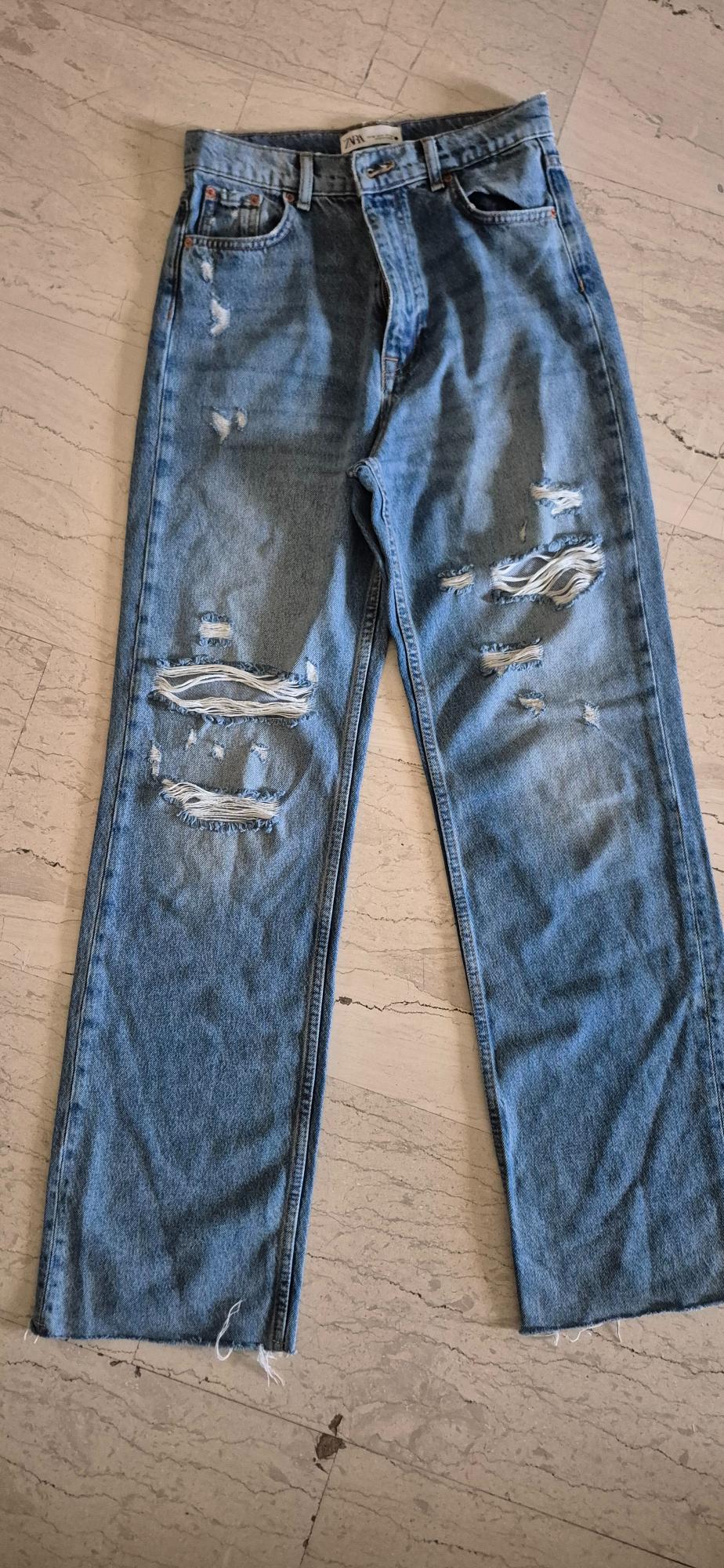Worn blue jeans with rips in the legs, on a light tiled floor. Visible label on the waistband.