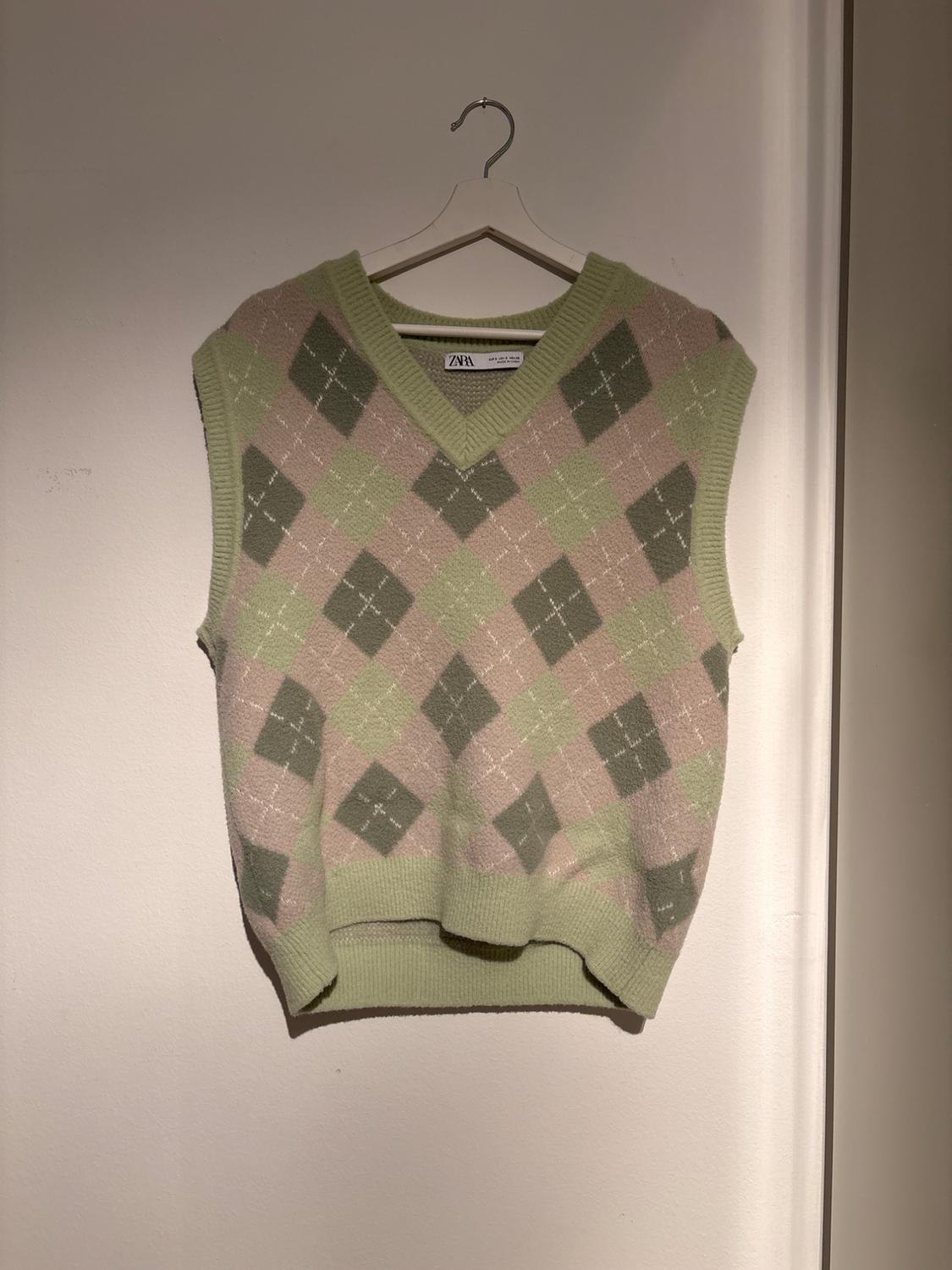 Sleeveless vest with a diamond pattern in green and pink tones, hanging on a white hanger against a light wall.