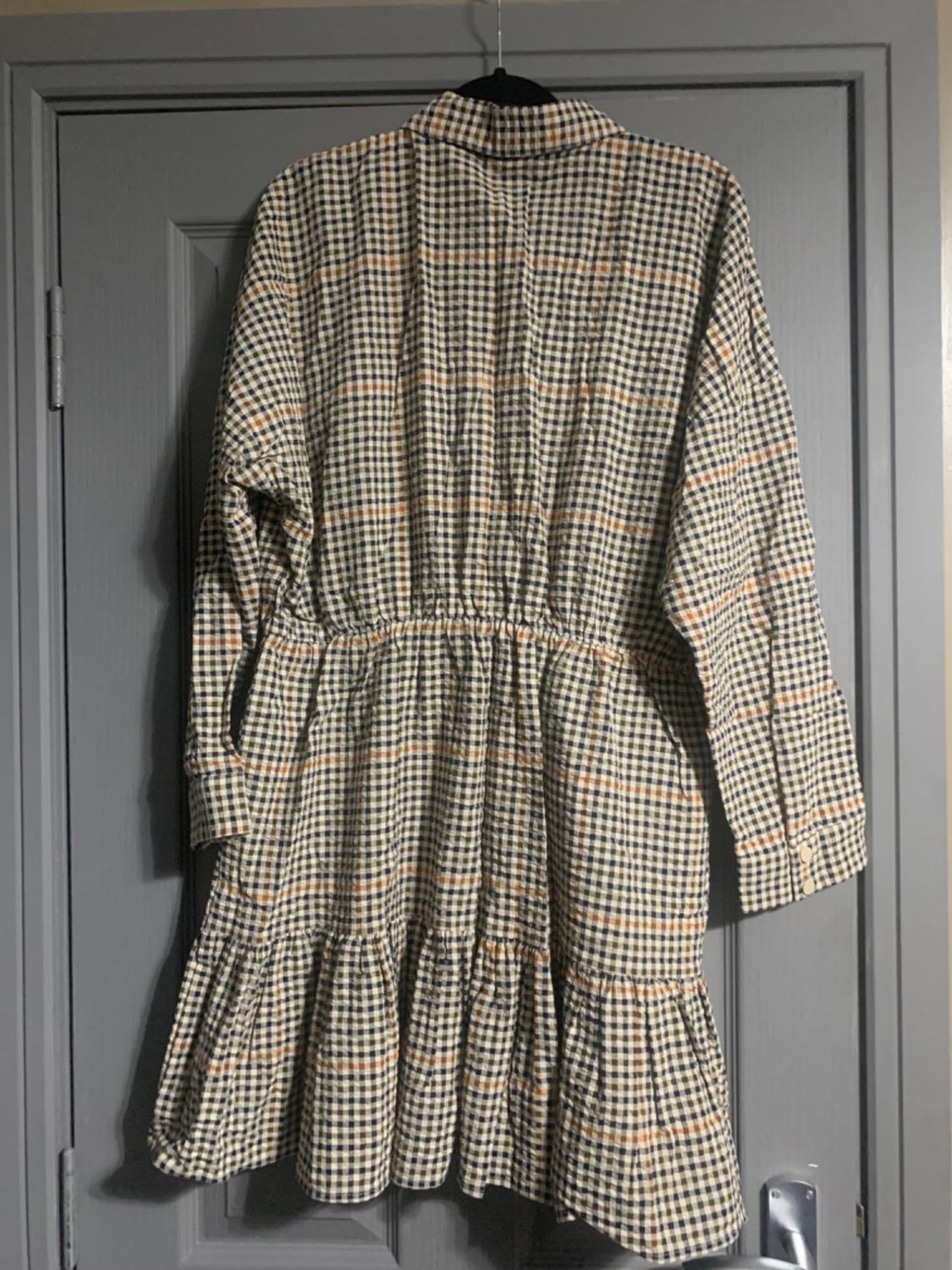 Checked dress in beige and black tones hanging on a grey door. Design with elasticated waist and ruffles at the bottom.