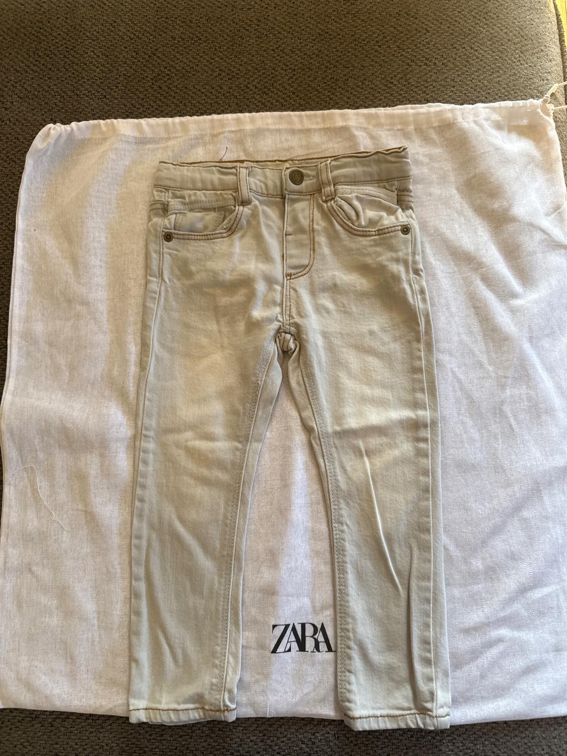 Beige children's trousers on a white bag with the Zara logo, placed on a brown textured surface.