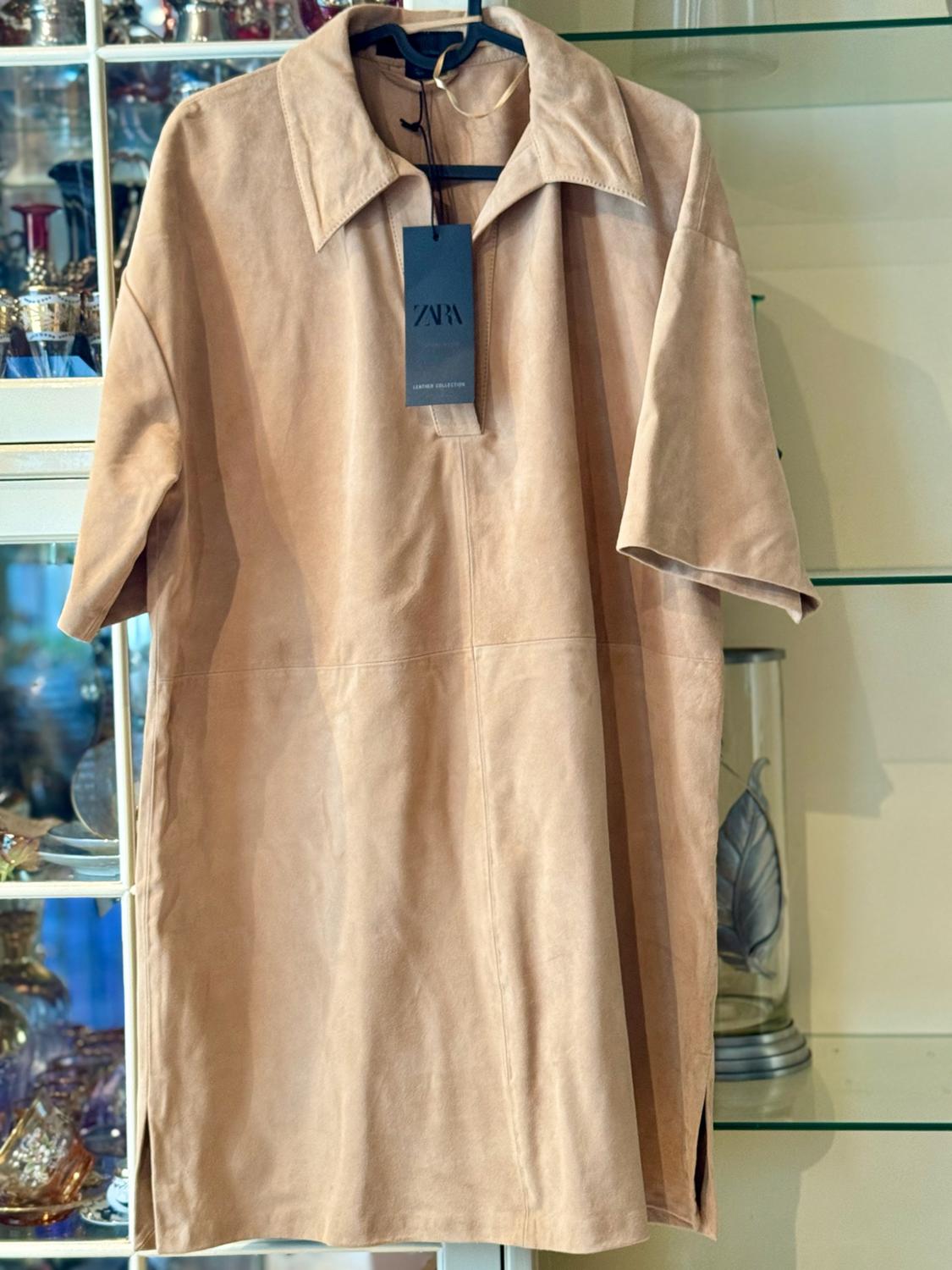 Long beige shirt with a collar and short sleeves hanging in front of a shop window. Black label with the text ZARA LEATHER COLLECTION.
