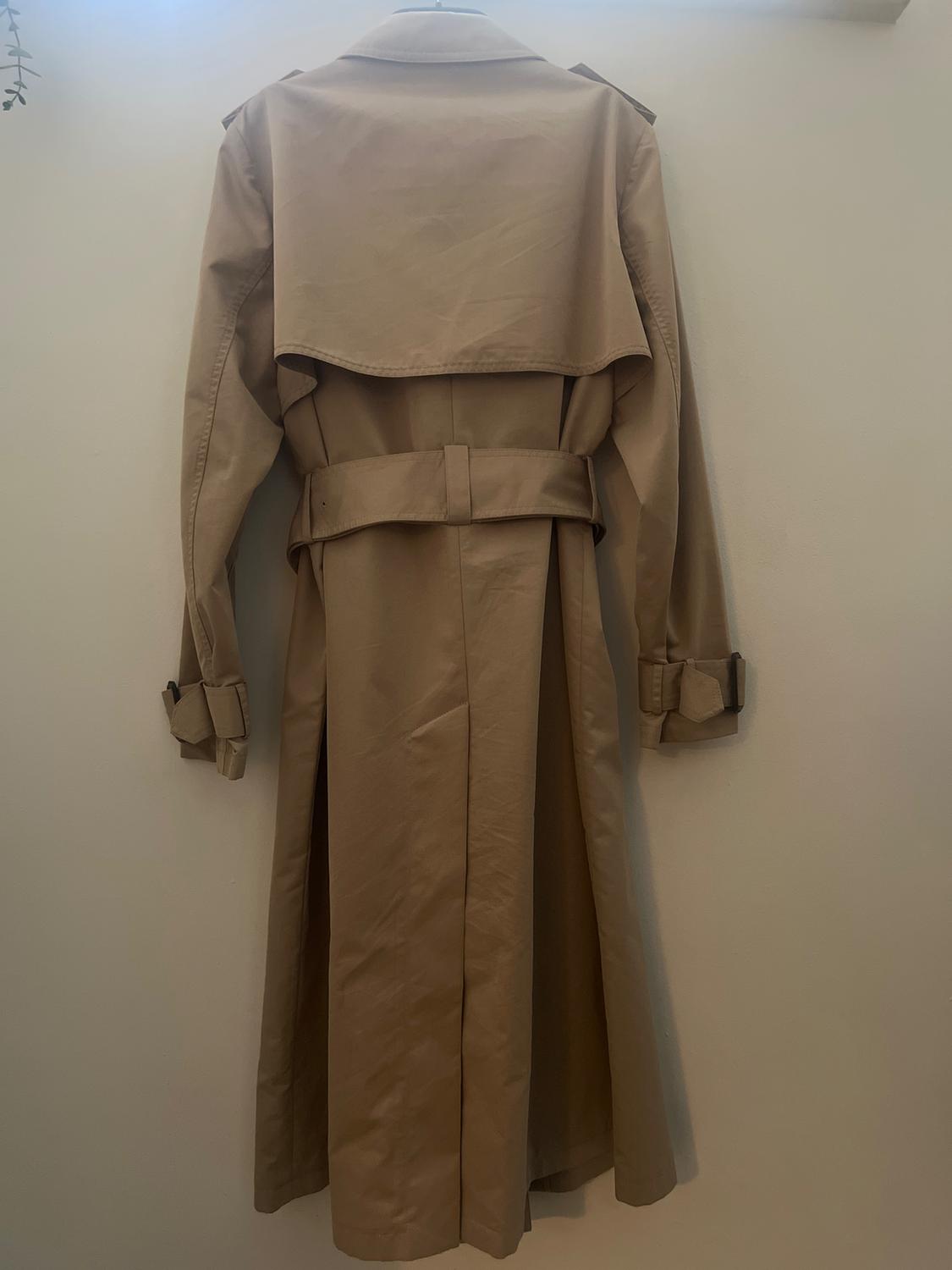 Beige trench coat hanging on a white wall, viewed from behind. It has a belt and detailing on the sleeves.