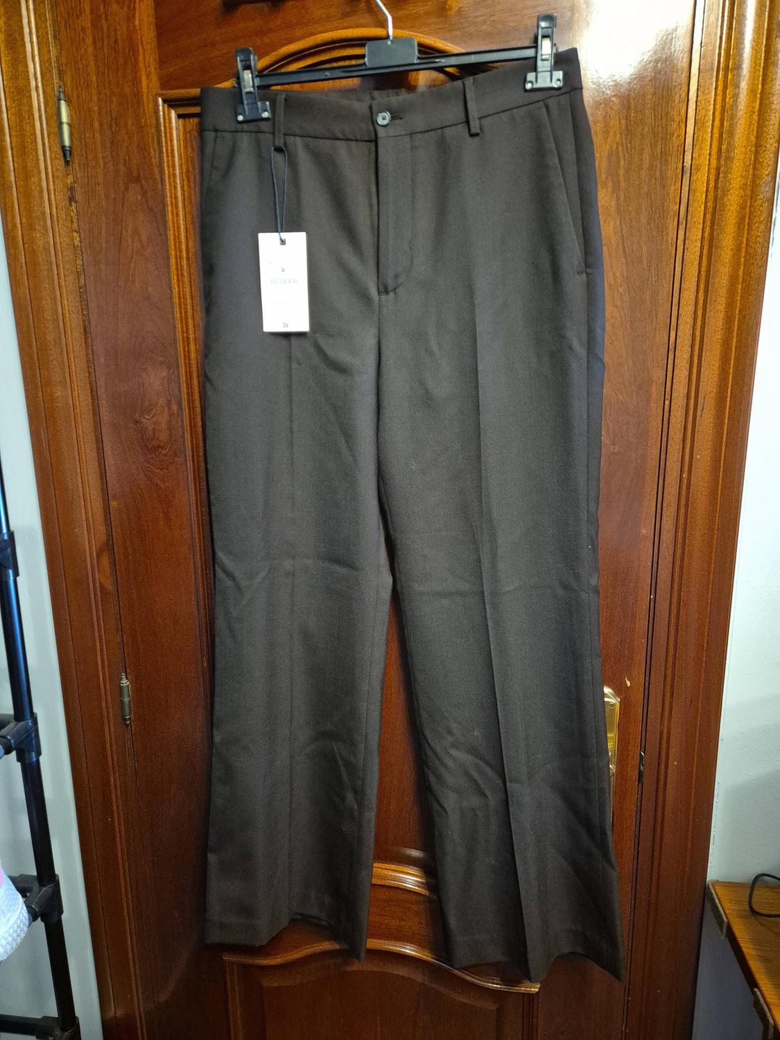 Dark brown dress trousers hanging from a hanger on a wooden door. They feature a front crease and a white label bearing the text “38”.