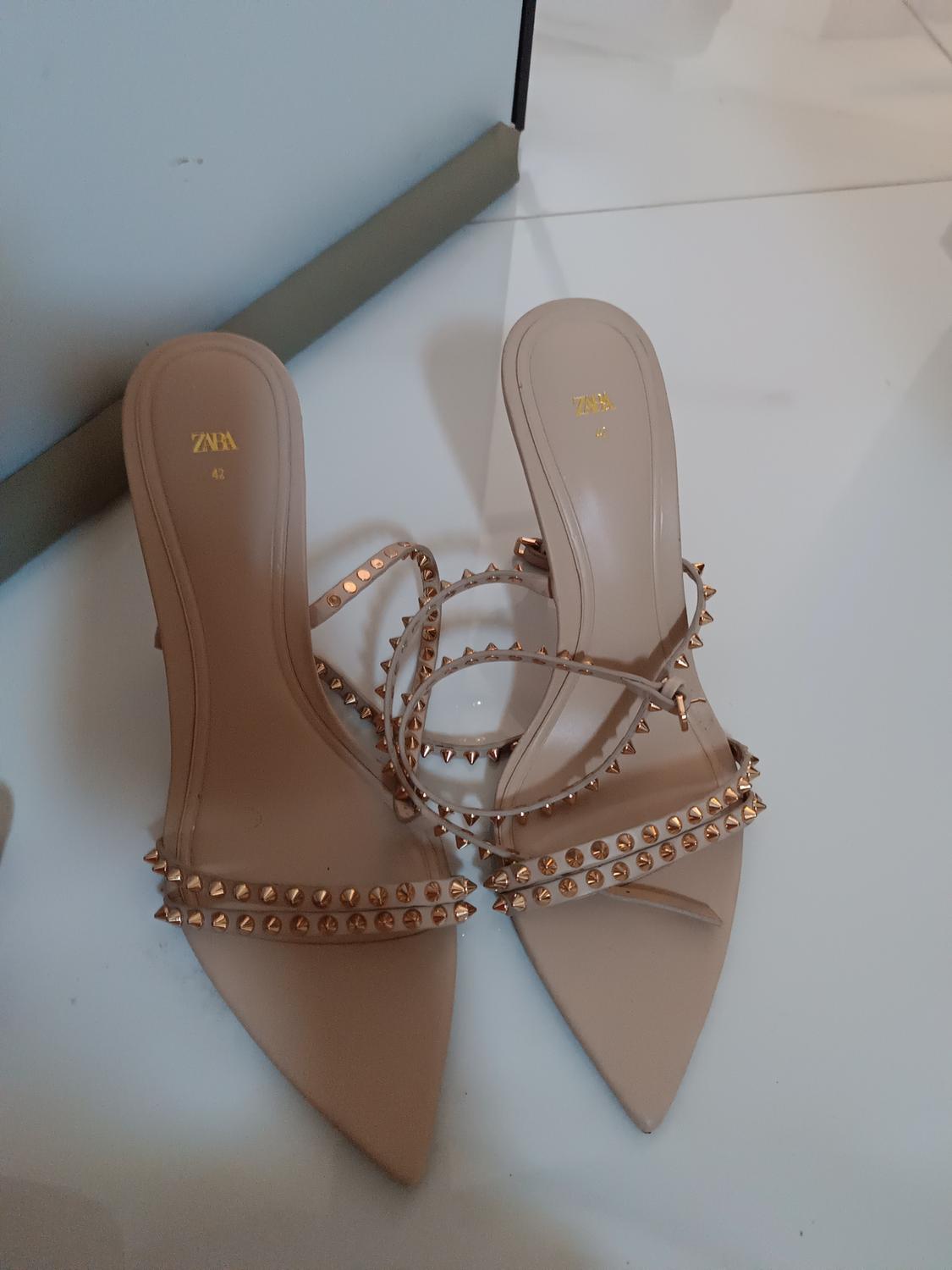 Beige heeled shoes with straps adorned with gold studs. ZARA brand visible on the insole. White sole.