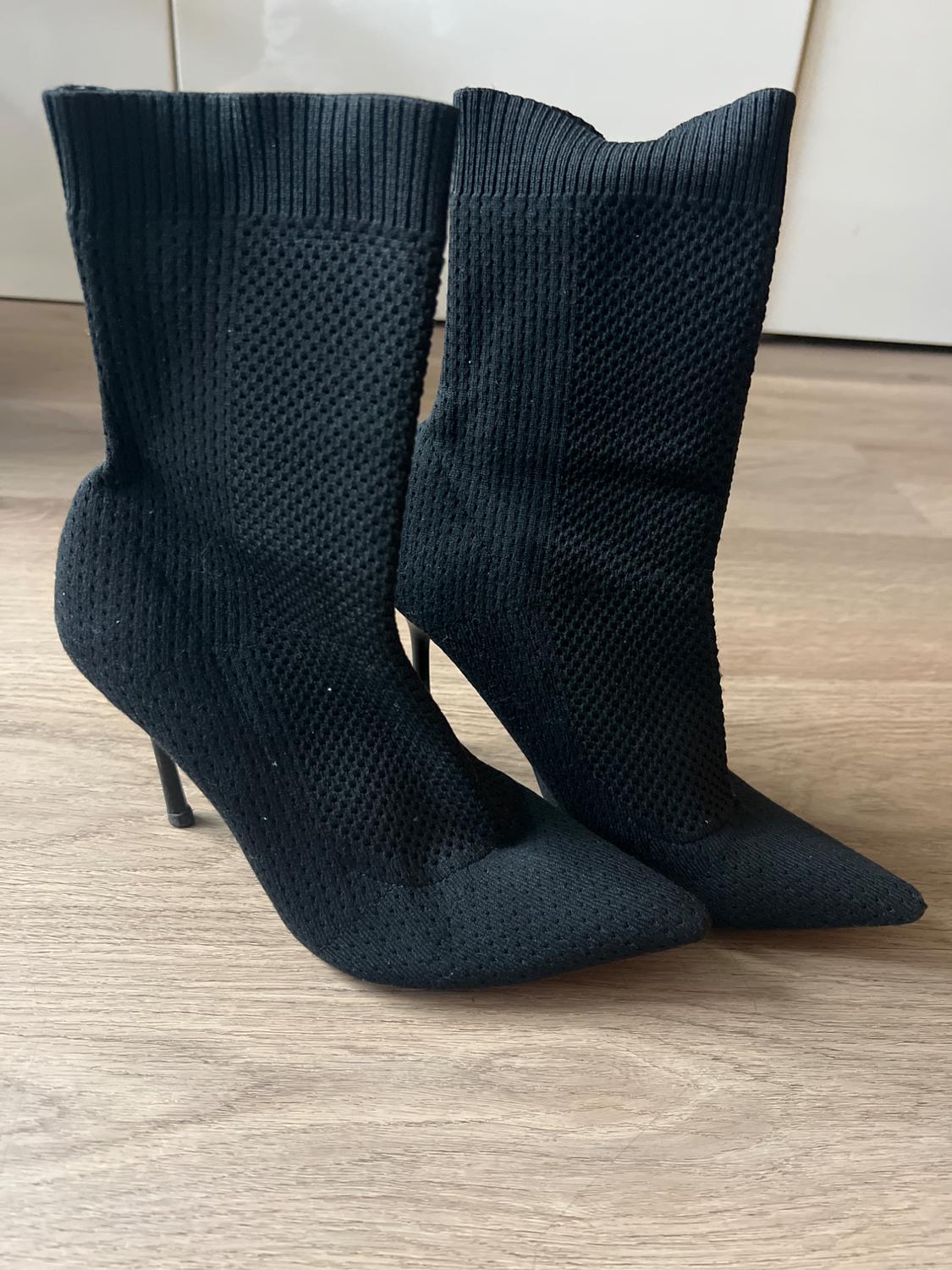 Pair of black knit sock-style ankle boots with stiletto heels and pointed toes, set on a light wood floor.