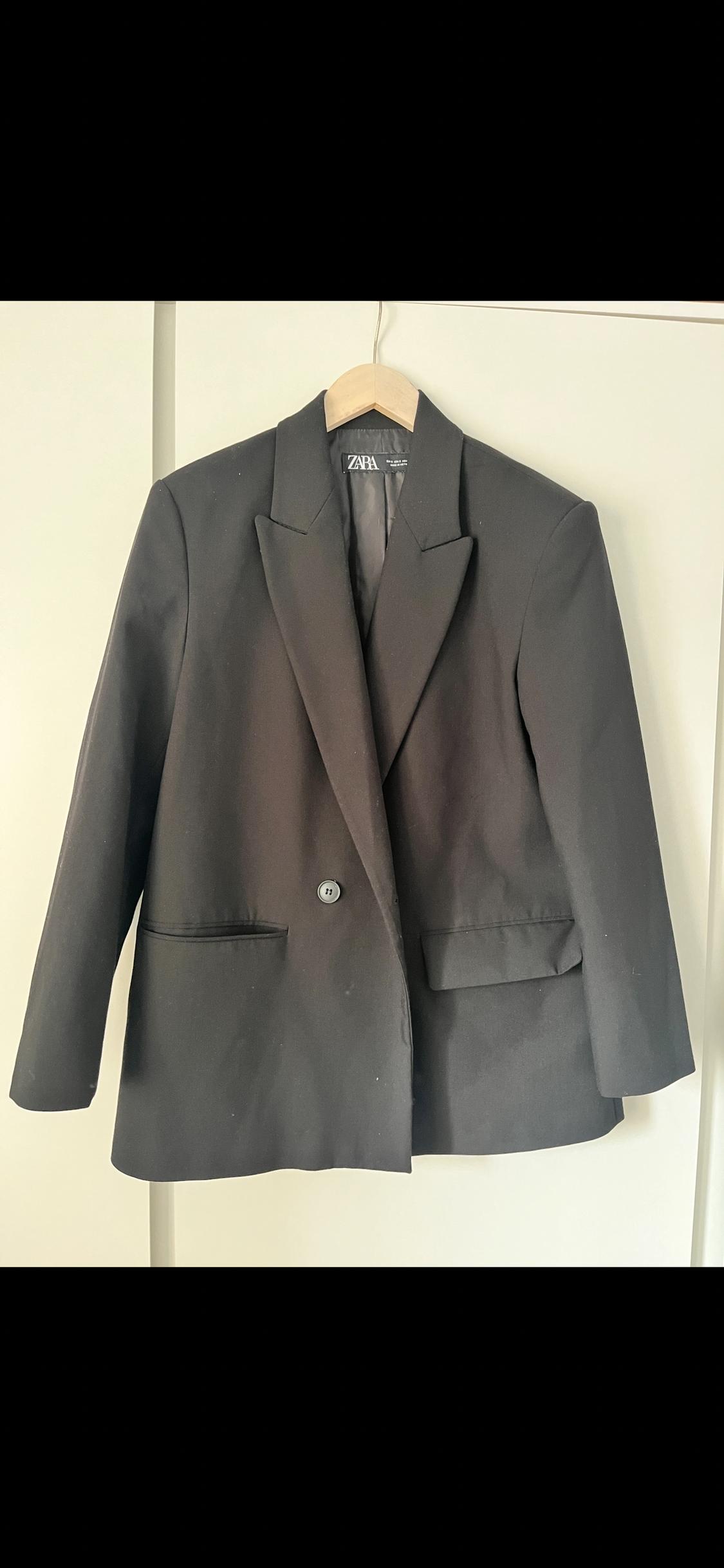 Black, single-button jacket hanging on a wooden hanger. Visible label with text "ZAPA" on the inside of the collar.