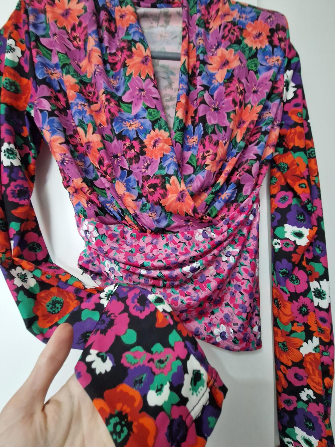Long-sleeved blouse with a striking multi-coloured floral pattern, wrap-over V-neck, and draped detail. A hand is shown holding the sleeve against a white background.