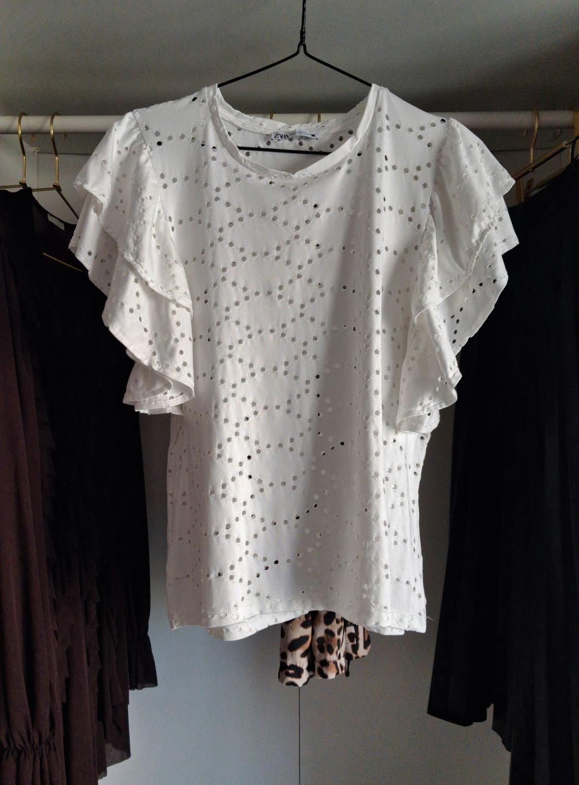 White blouse with ruffles and a perforated design hanging on a clothes rail, surrounded by dark garments. Visible tag: "Zara".