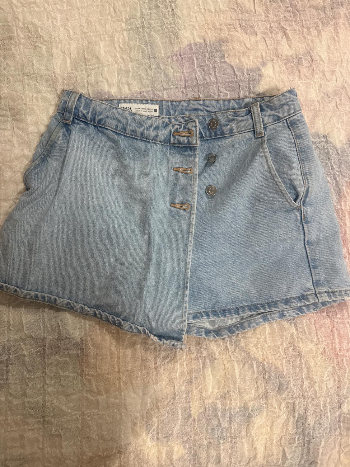 Light blue casual denim skort with asymmetrical buttons, on a textured bedspread. ZARA label.