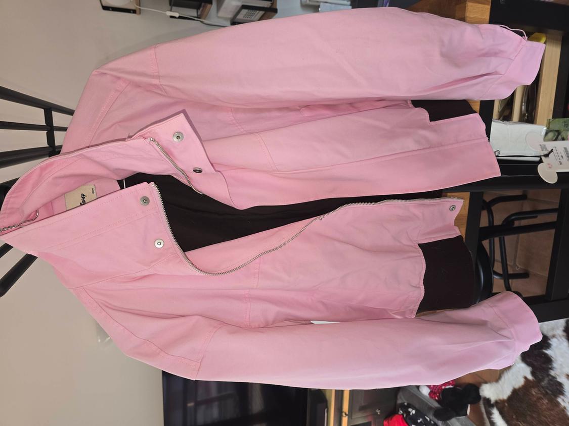 Pink jacket hanging on a black clothes rail, with a zip and silver buttons. Modern interior background with furniture and decor.