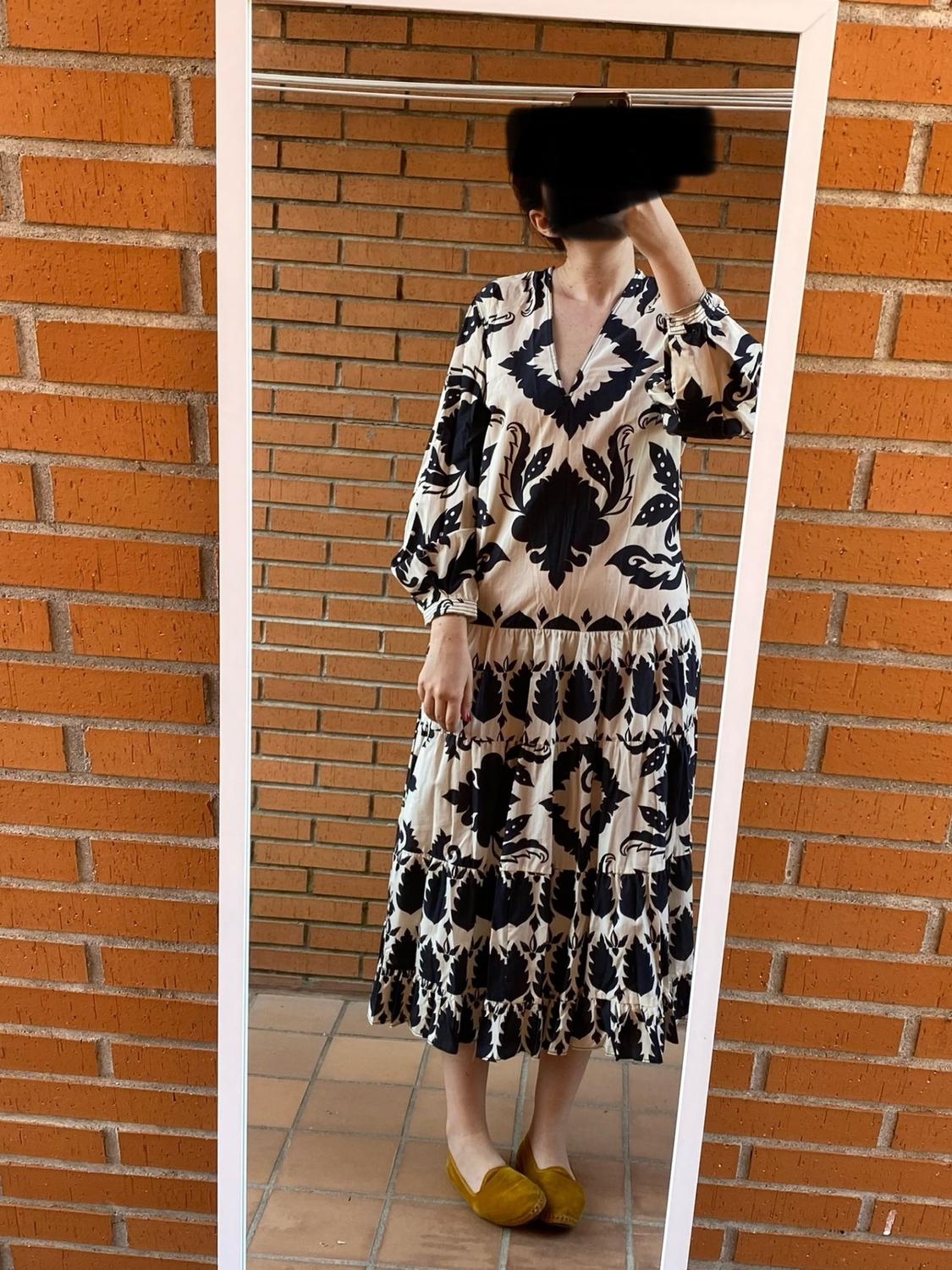Woman in front of a mirror wearing a black and white patterned dress, yellow shoes, with a brick wall background.