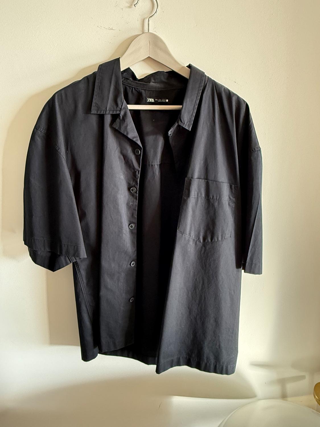 Black short-sleeved shirt hanging on a wooden hanger against a white wall. Visible label with text "Zara".