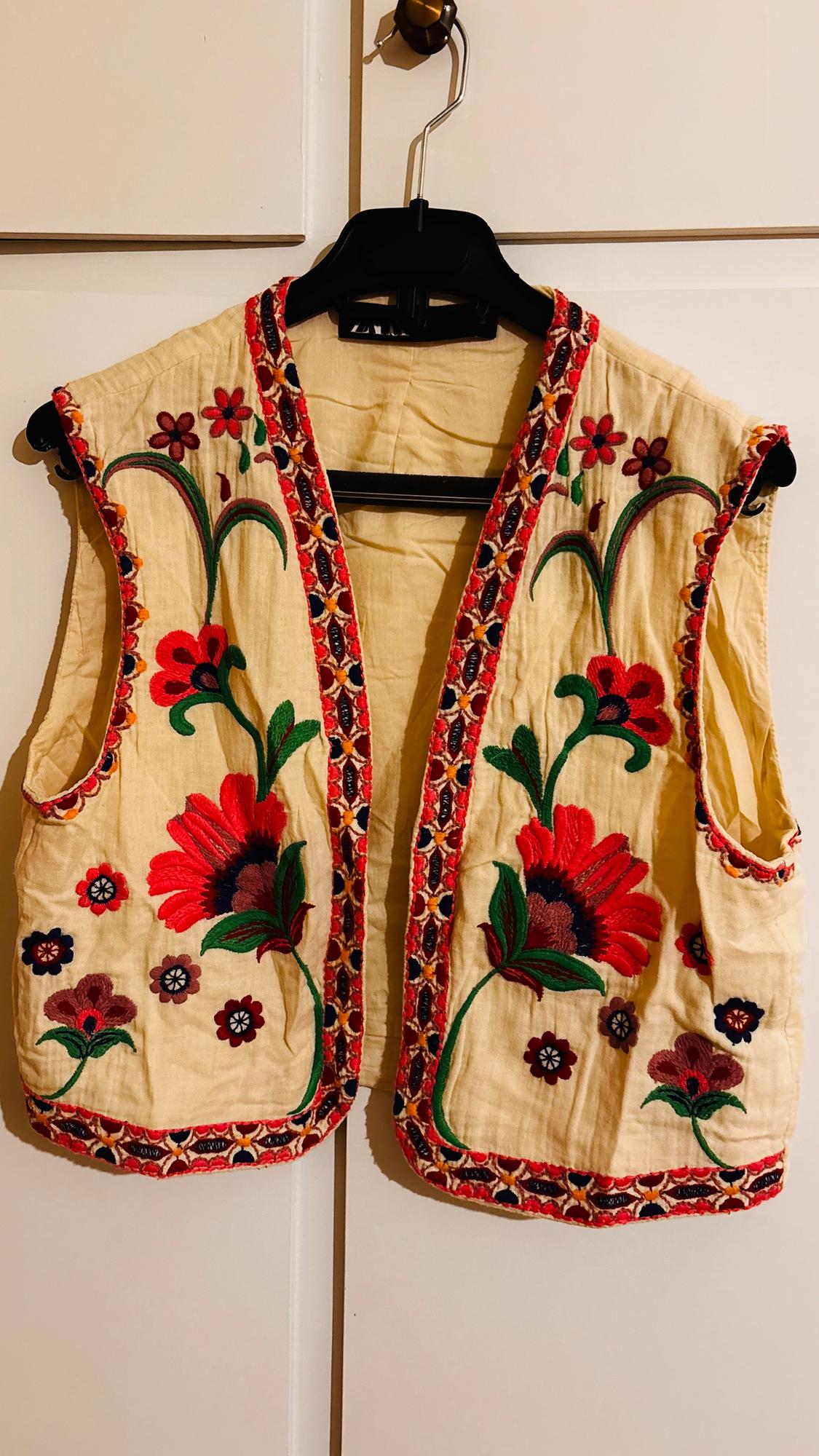 Beige waistcoat hanging on a hanger, with floral embroidery in red, green, and brown, and decorative red and orange trim.