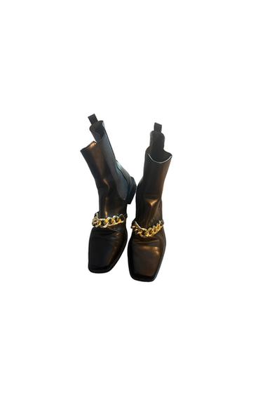 Black square-toe booties with elastic side panels and gold chain detail on the instep, set against a white background.