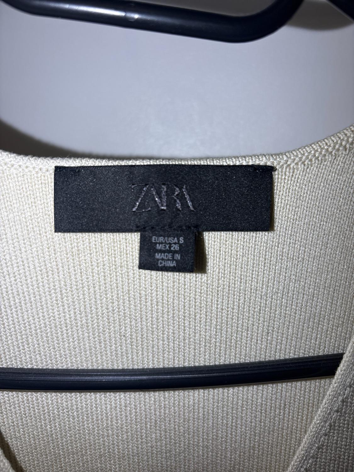 Black label on a cream-coloured knit garment hanging on a hanger. Text: ZARA EUR/USA S MEX 26 MADE IN CHINA.