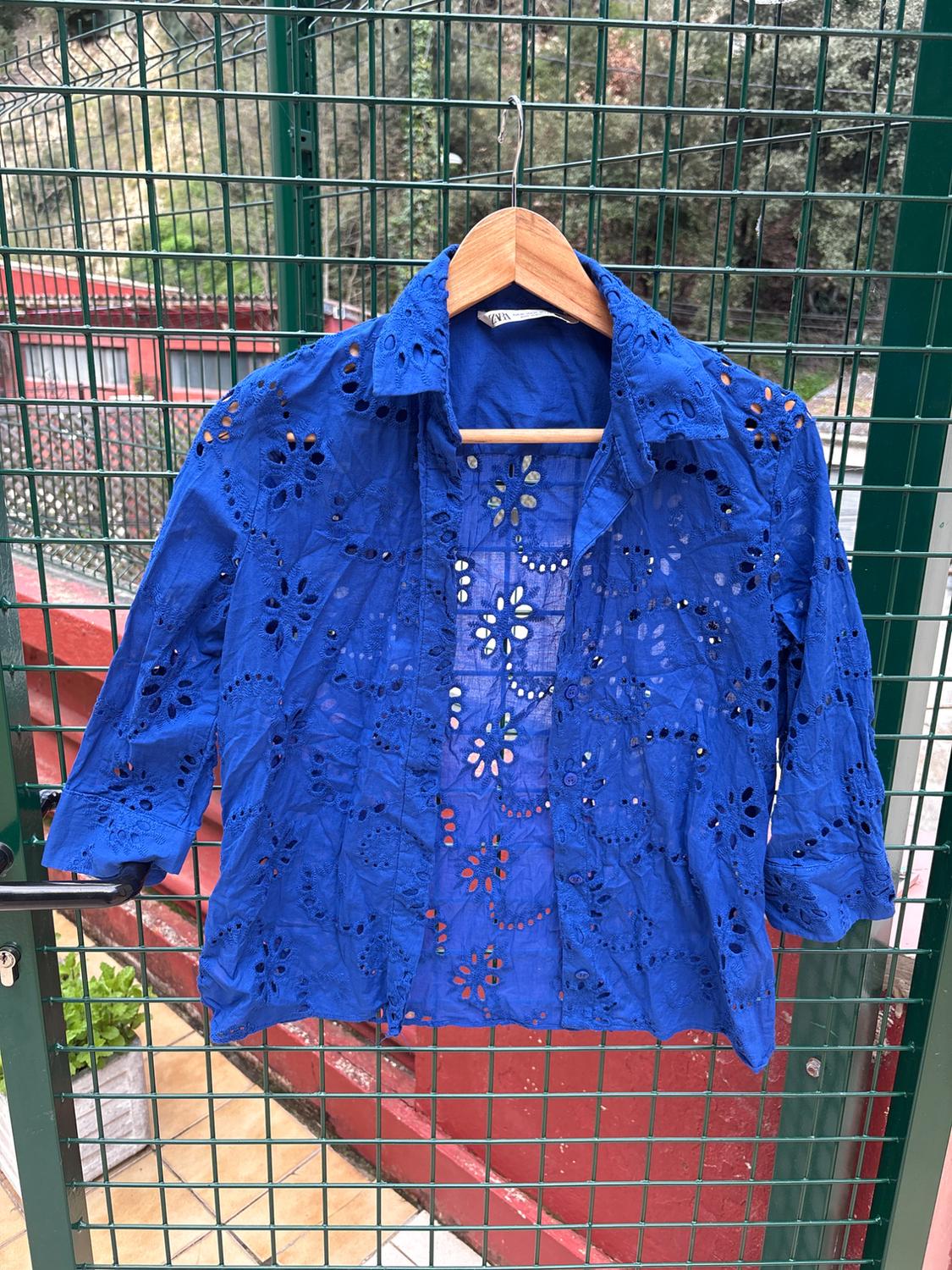 Blue blouse with floral cut-out design hanging on a wooden hanger over a metal grid, garden and red building in the background.