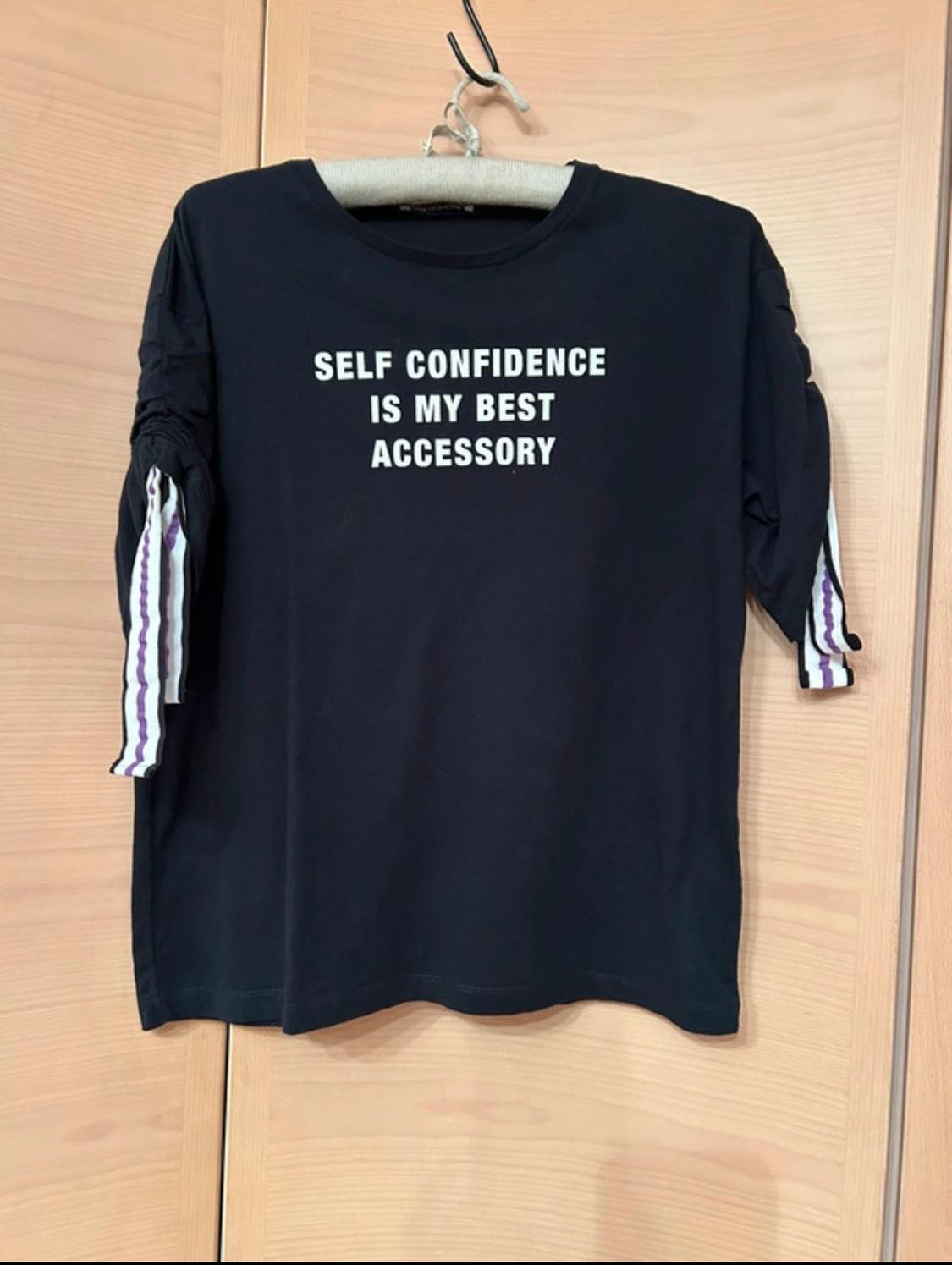 Black t-shirt hanging with "SELF CONFIDENCE IS MY BEST ACCESSORY" text in white. Sleeves with purple and white stripe details.