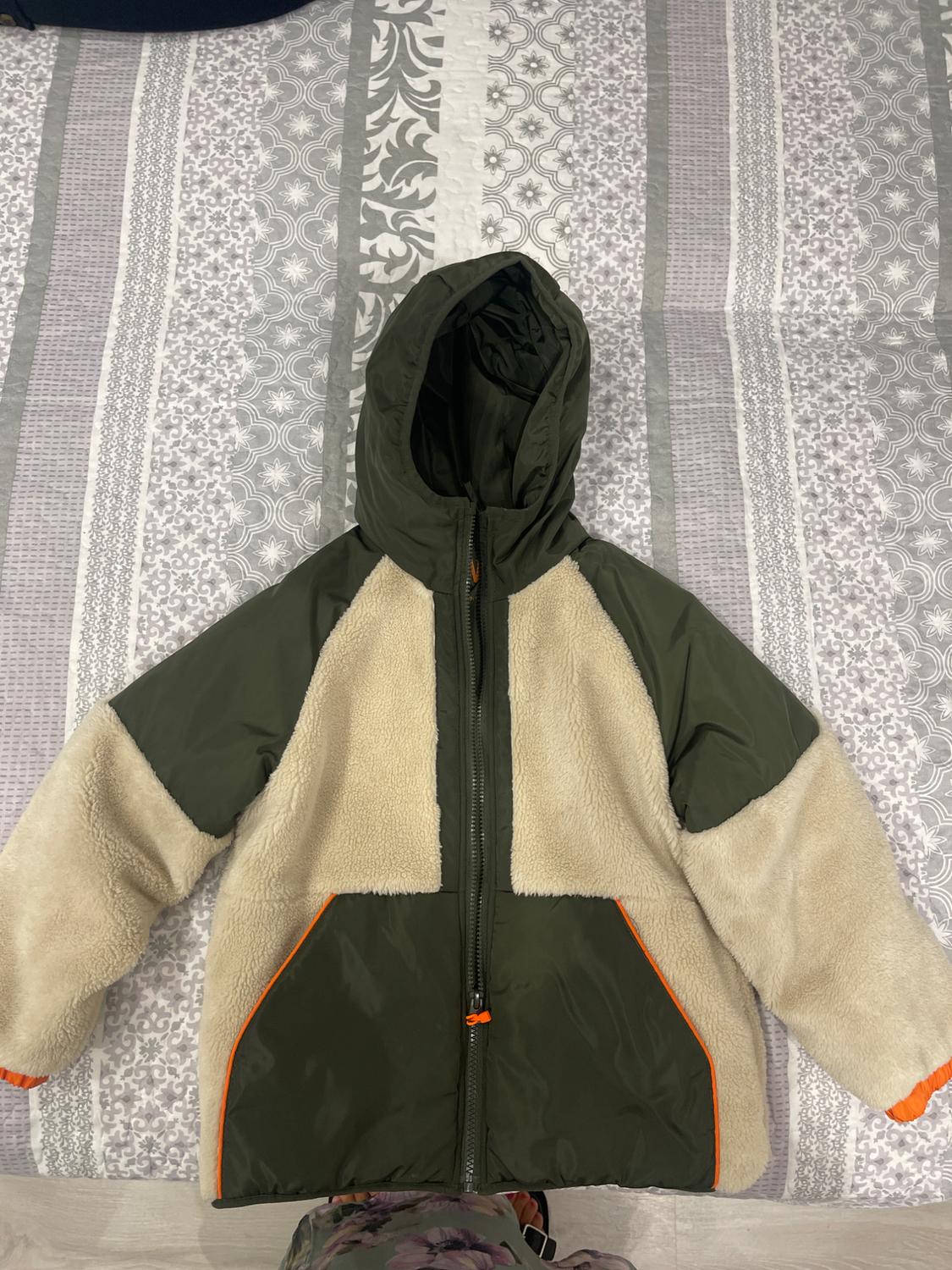 Green and beige hooded jacket over a patterned quilt. Orange details on the pockets and cuffs.