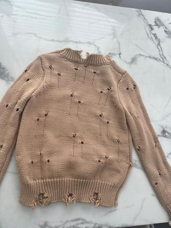 Beige knit sweater with decorative holes and frayed edges, on a white marble surface with gray veins.