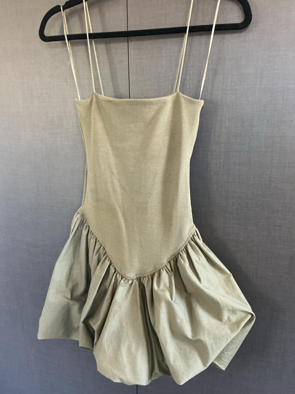 Olive green spaghetti strap dress, fitted V-neck bodice, and voluminous short skirt, hanging on a black hanger against a gray background.