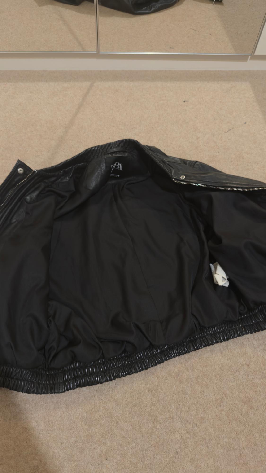 Black jacket open on a beige rug, reflected in a mirror. Visible label with "ZARA" lettering. Relaxed and modern atmosphere.