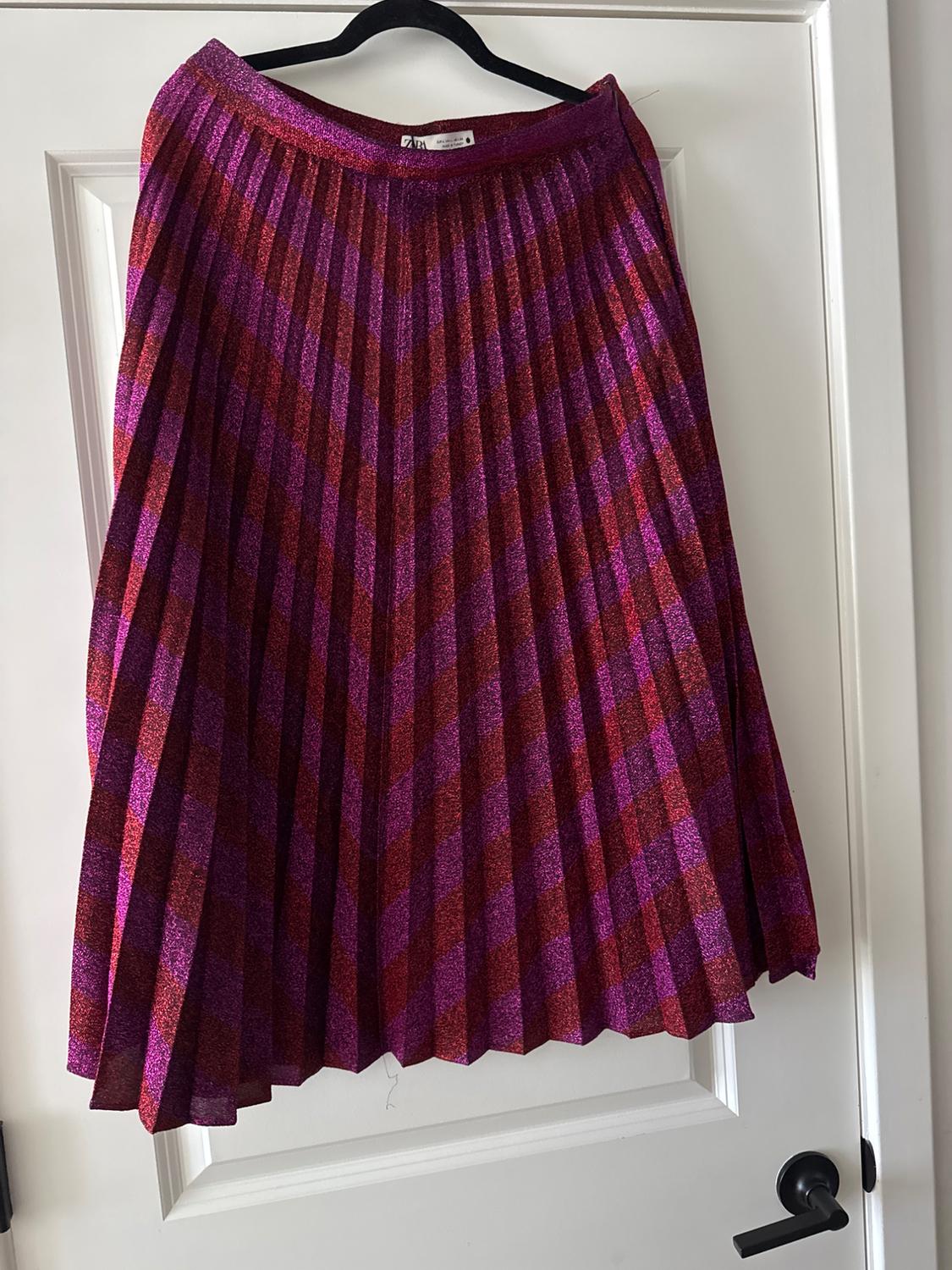 Pleated skirt in shades of purple and red with a diagonal stripe pattern, hanging on a black hanger over a white door.