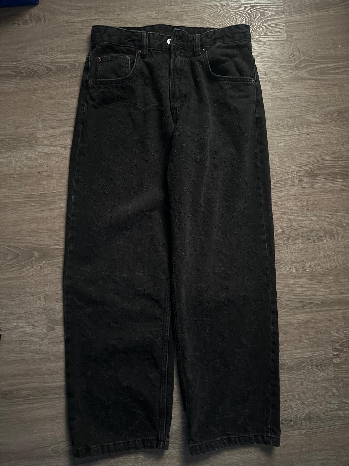 Black denim pants with a subtle design, placed on a light wooden floor.