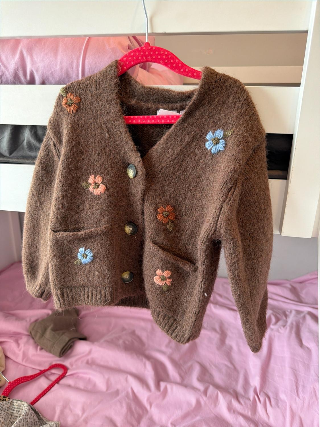 Brown knitted cardigan with embroidered flowers, hanging from a pink hanger on a white bunk bed above a bed with pink sheets.