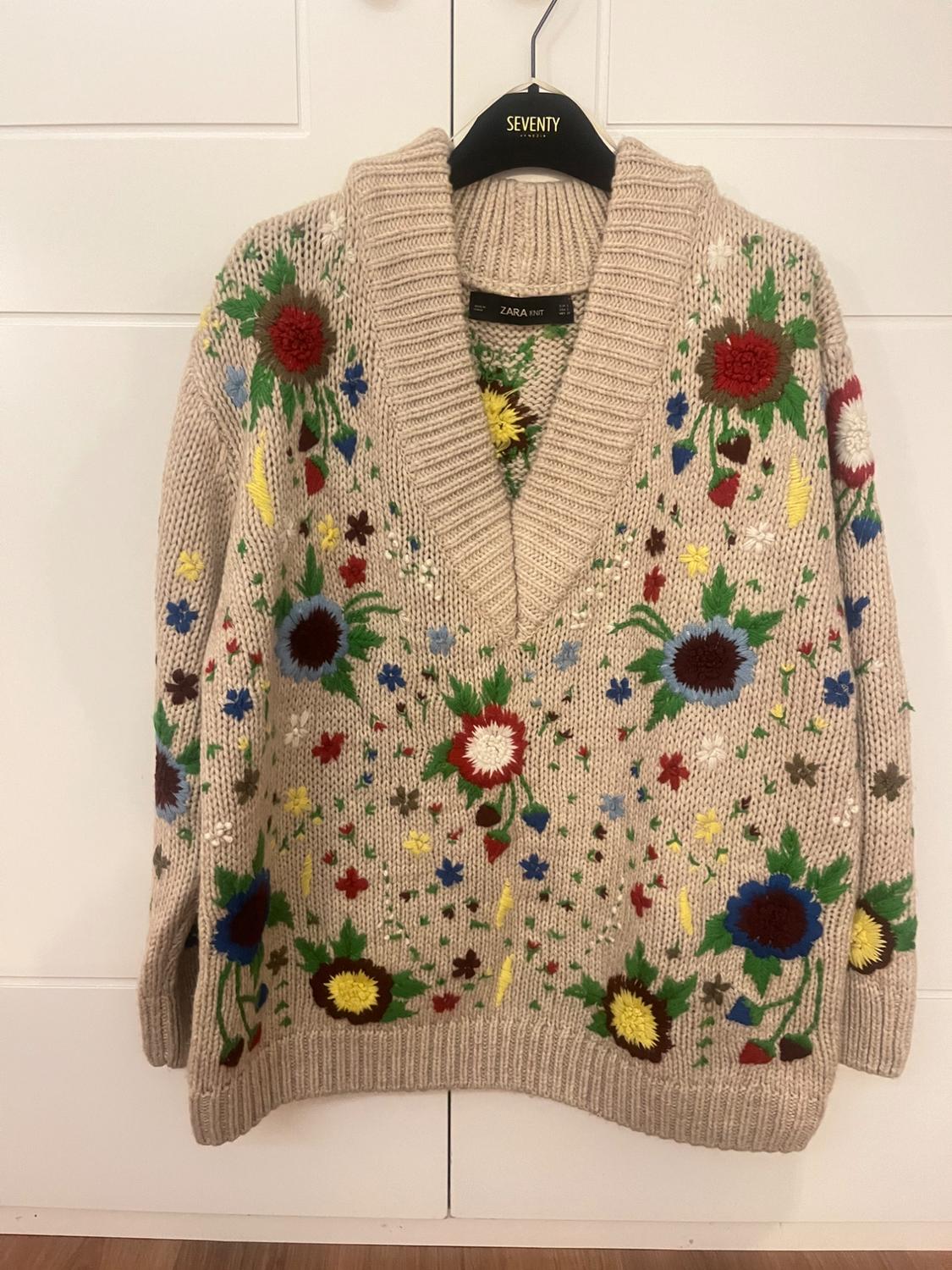Beige jumper with embroidered flowers in red, blue, yellow, and green. Hanging on a black hanger with the brand "SEVENTY".
