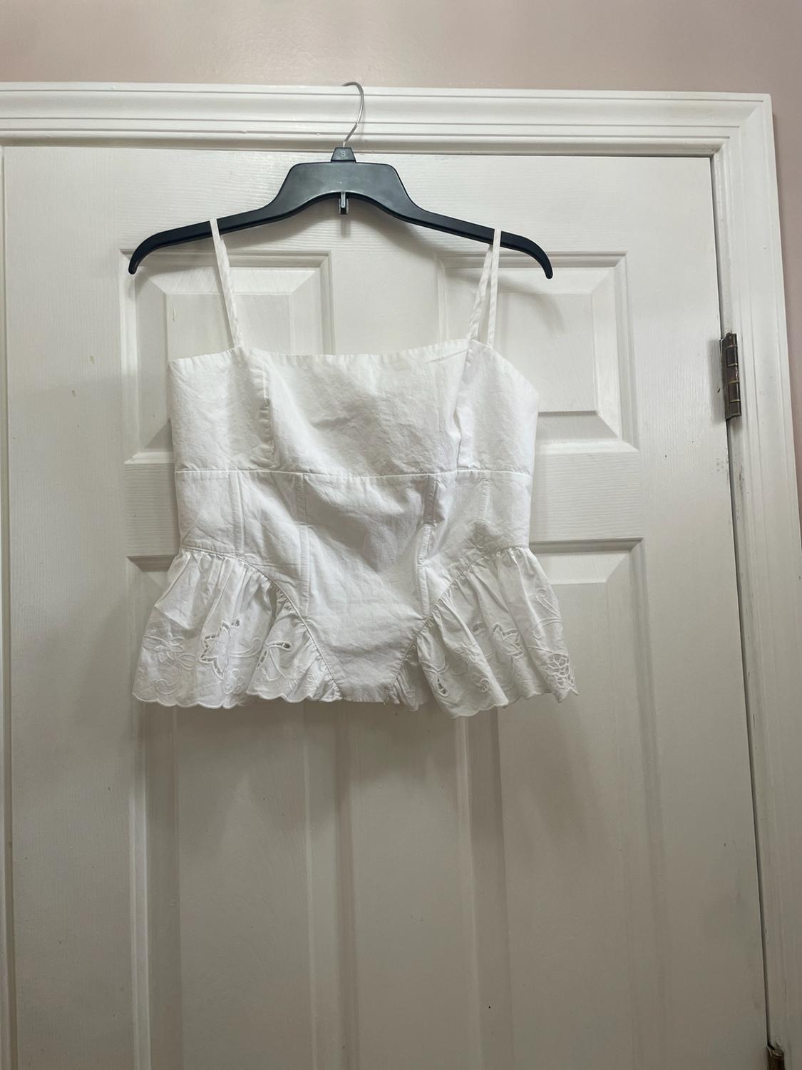 White camisole with thin straps, a square neckline, and an embroidered ruffle hem, hanging on a black hanger over a white door.