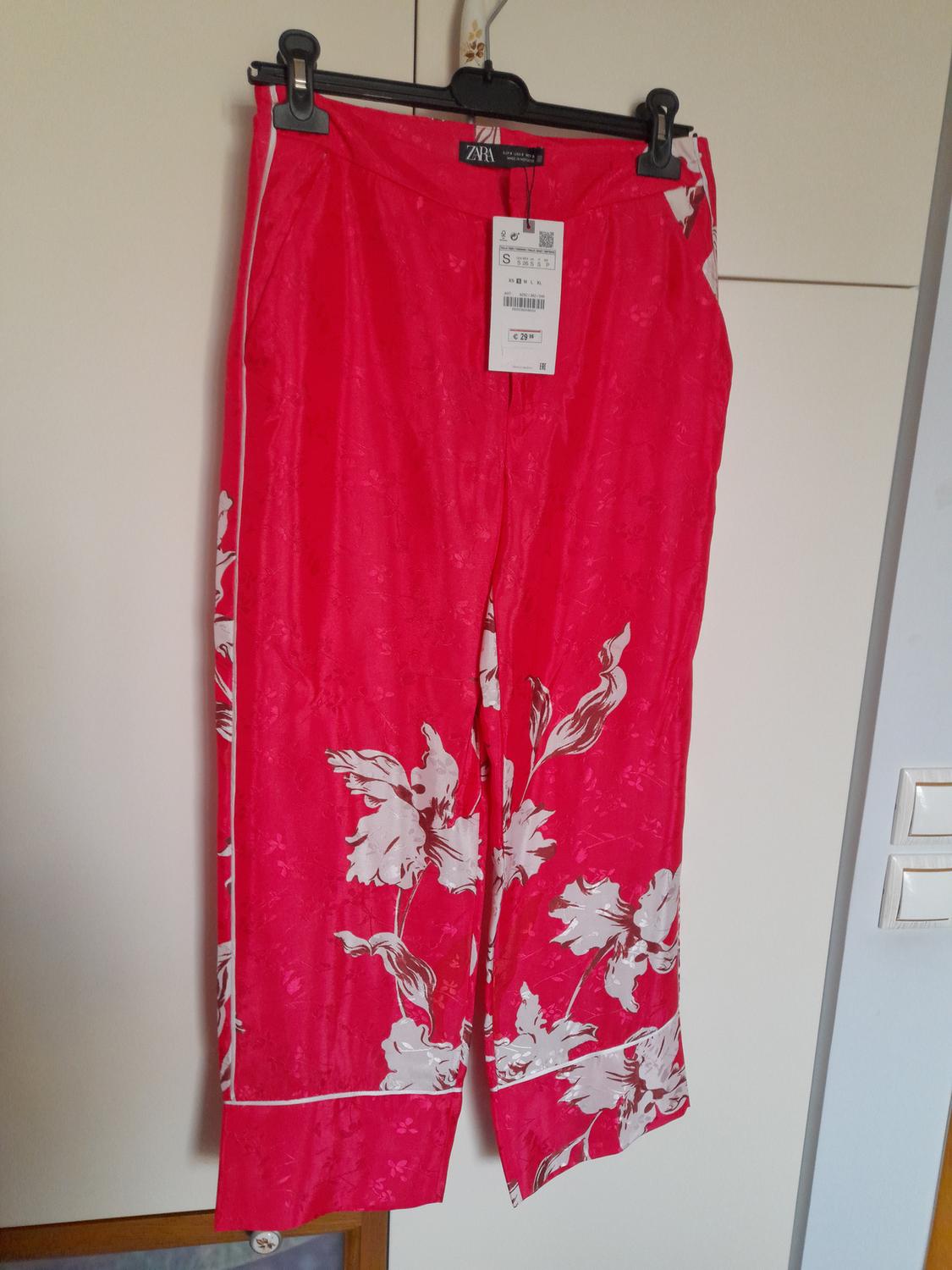 Red trousers with a white floral print, hanging on a hanger. Visible tag with a price of €29.95. White wardrobe background.