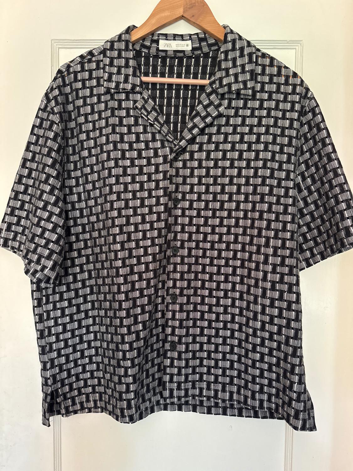 Short-sleeved ZARA shirt with a black and white geometric print, buttoned up and hanging on a wooden hanger on a white door.