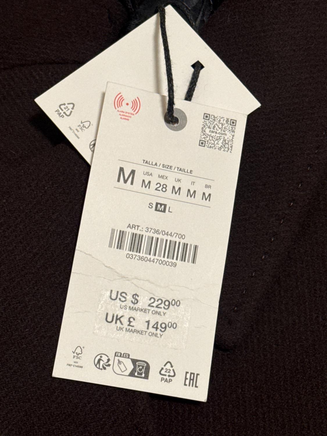 Clothing tag with price in dollars and pounds, size M, barcode and QR. Text: "US $229, UK £149, US MARKET ONLY, UK MARKET ONLY".