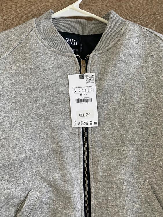 Gray jacket with zipper and $89.00 price tag hanging on a white hanger against a wooden background.