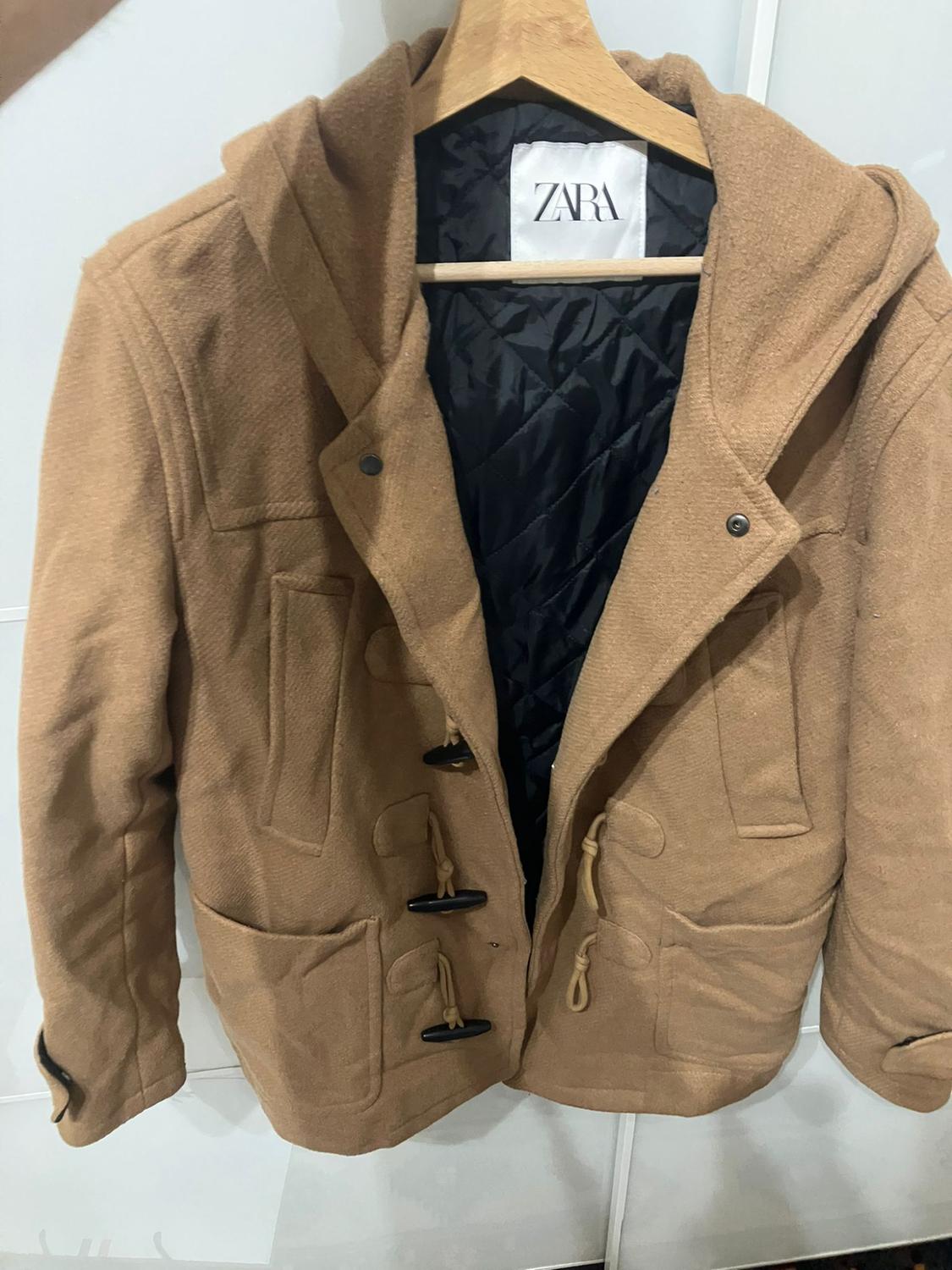 Brown coat with a hood and toggle fastening, hanging on a hanger. Visible black padded interior. "ZARA" label on the collar.