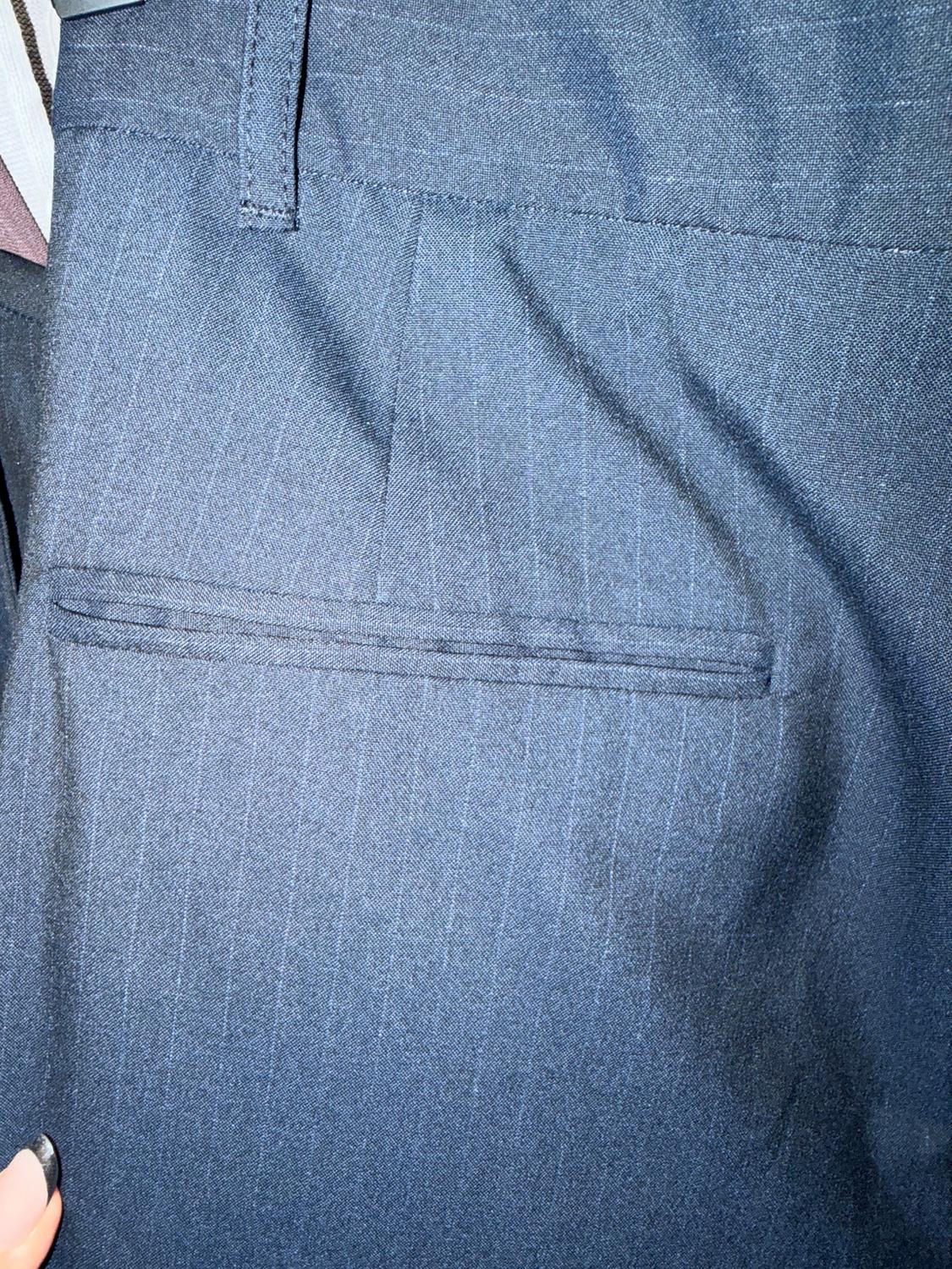 A close-up of dark blue pants with a back pocket and thin vertical stripes. A hand with black painted nails holds the edge.