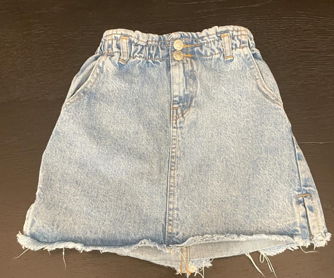 Light blue denim skirt with an elasticated waist, two front buttons, pockets and a frayed hem, on a dark wood background.