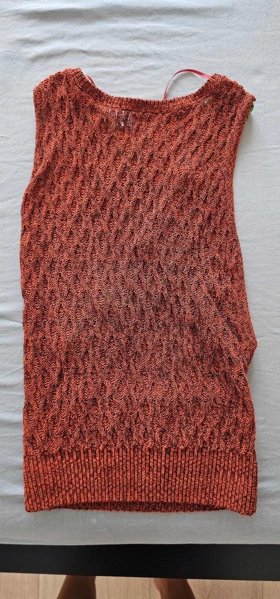 Sleeveless openwork knitted top in terracotta with a ribbed hem, laid out on a light grey fabric.