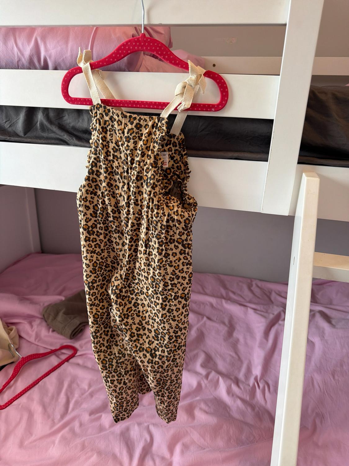 Baby's leopard print jumpsuit with bow straps, hanging on a pink hanger over a white bunk bed.