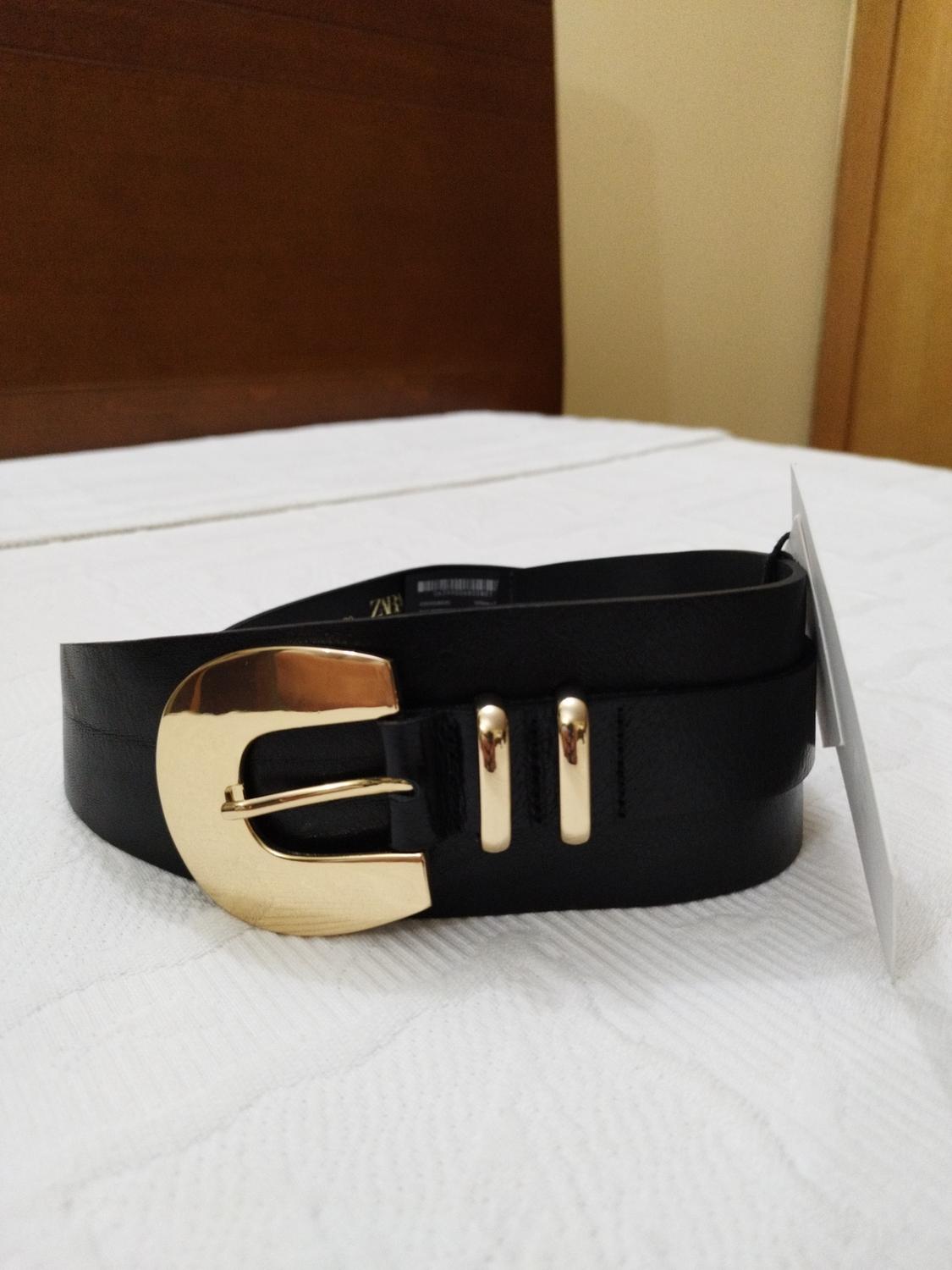Black belt with gold buckle on a white bed. Brown wooden background and beige wall. Visible tag on the belt.