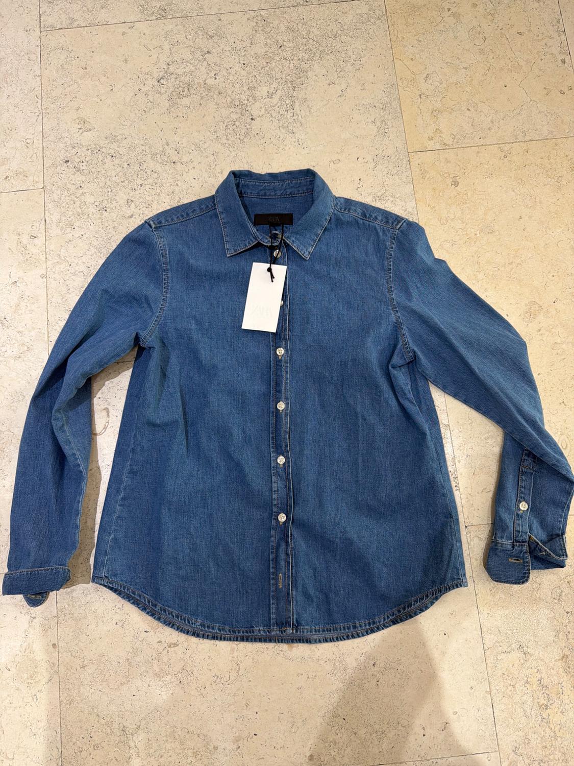 Blue long-sleeved button-down denim shirt with ZARA label, laid out on a light stone tiled floor in a relaxed style.