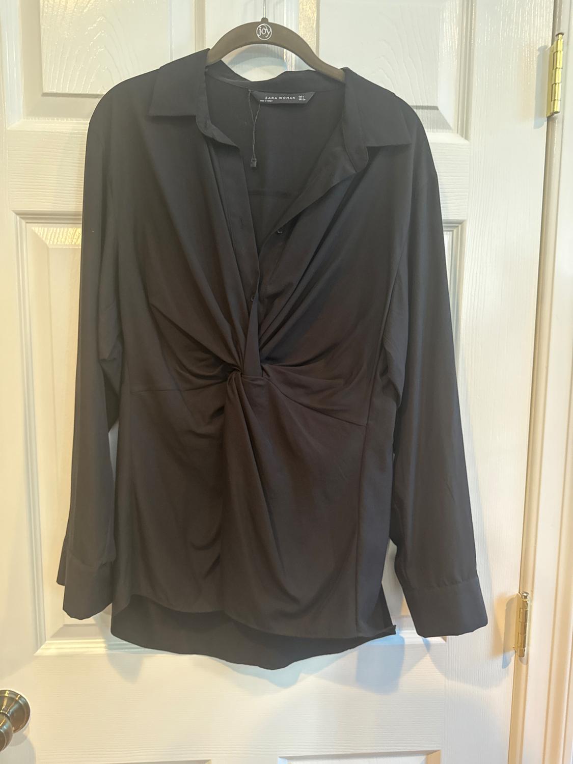 Black long-sleeved blouse with draped design on the front, hanging on a hanger over a white door.