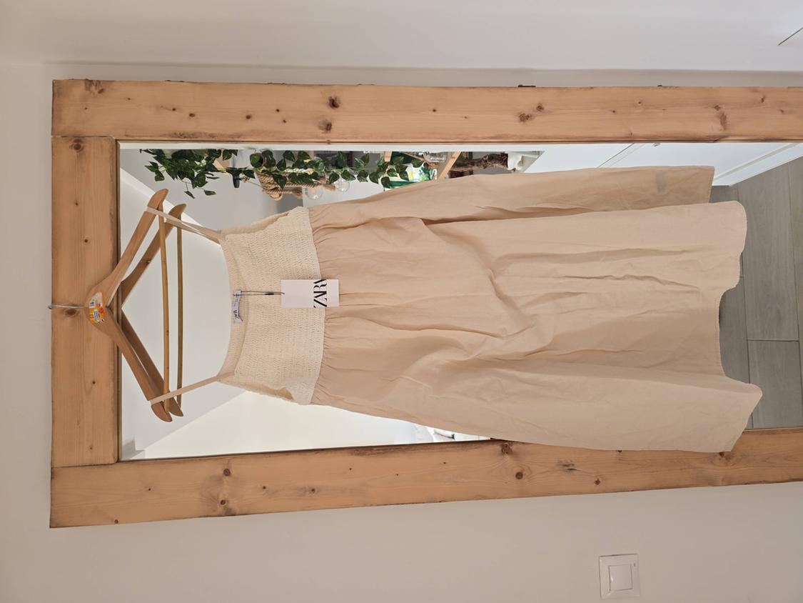 Beige dress hanging on a wooden hanger in front of a mirror with a wooden frame. Visible label with "ZARA" text.