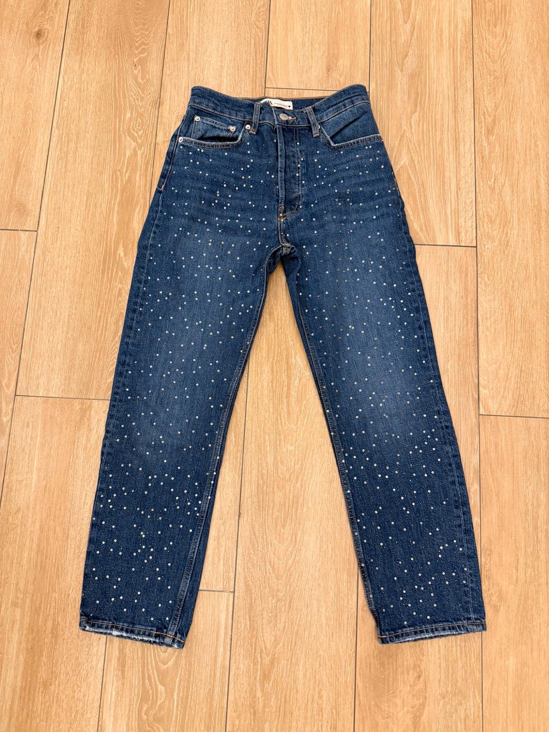 Blue denim trousers with sparkling rhinestone details, laid out on a light wooden floor.