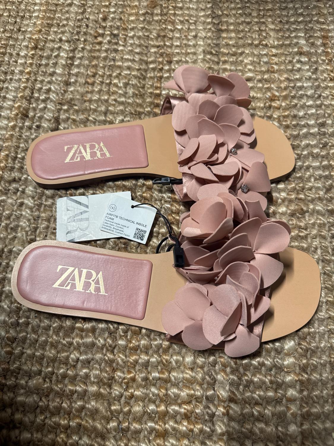 Pink flat sandals with floral embellishments and a "ZARA" logo on the insole, on a woven rug. Visible tags next to them.